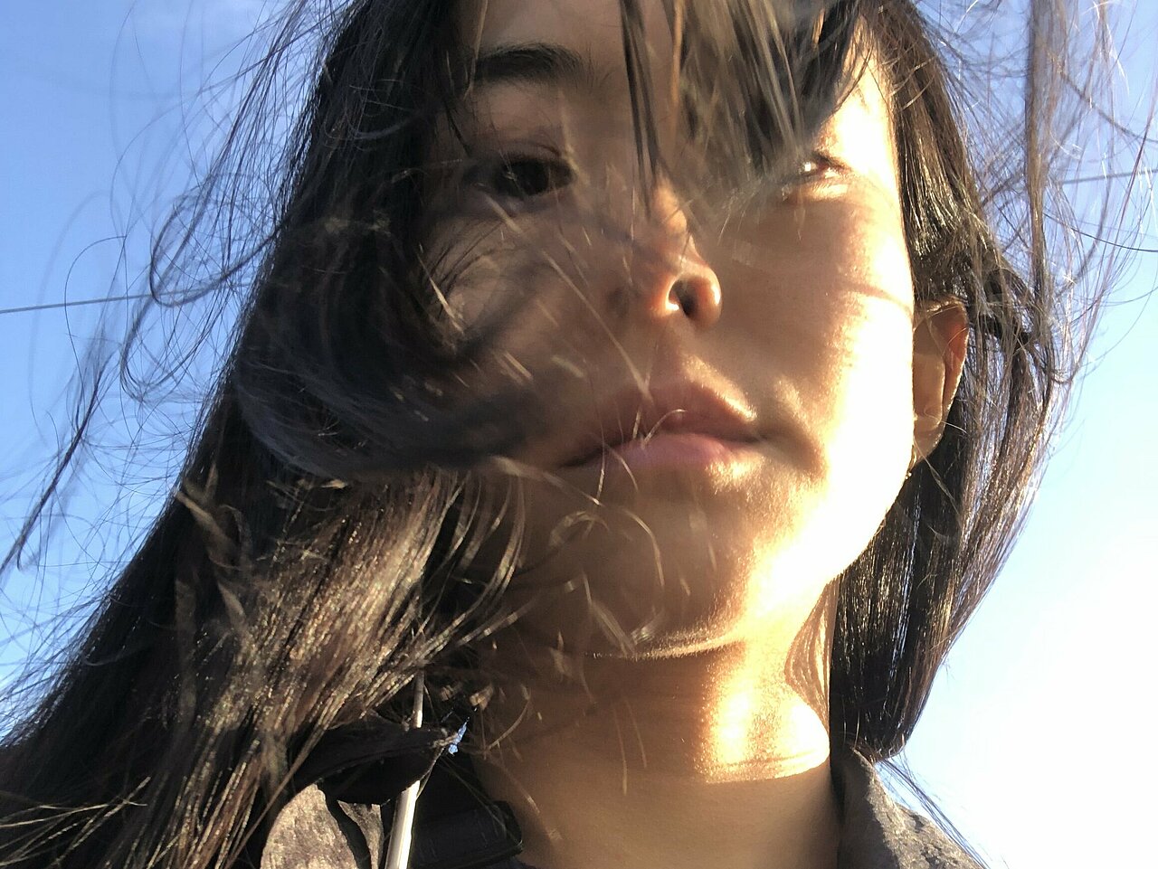 Portrait of Hanako Hayakawa, a selfie from below, hair being blown in the wind, blue sky in the background.