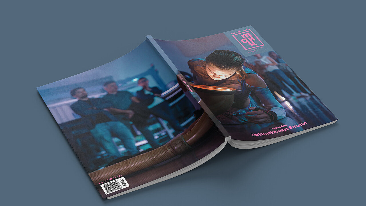 Magazin, with the opened pages turned towards the grey surface it is lying on. On the cover a dancer with her face very close, almost as if looking into a headlight. in the background, blurry, audience standing and watching.