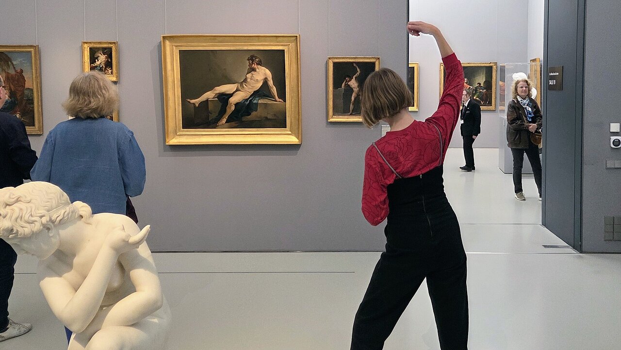 Eliza, seen from behind, standing in front of a painting, imitating the graceful posture