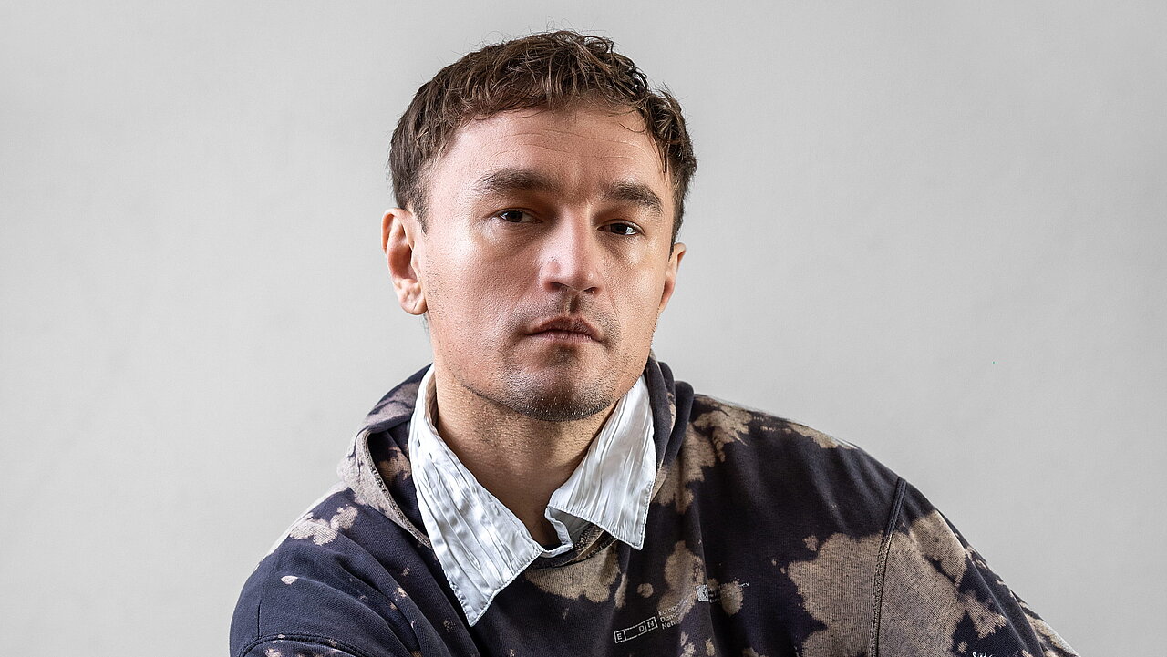 Portrait of Maciej Sado, sitting on a stool, one knee up, with the arms wrapped around. 