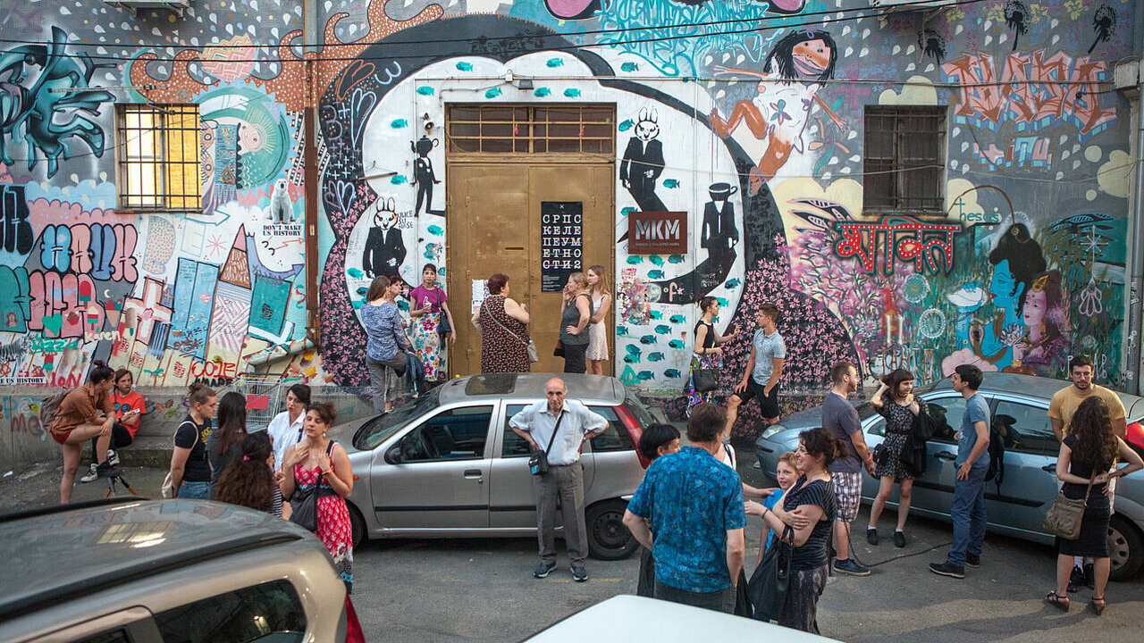 The golden entrance door of Magacin / Station Belgrade, in a wall covered in colorful grafitti, guest hangignn out in front of the building, between parking cars.