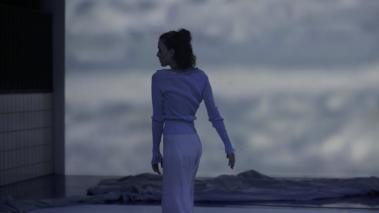 Jasmin standing in front of a big projection of the sea, swinging her arms.