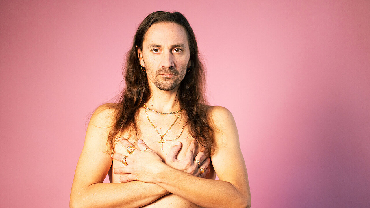 Luigi Guerrieri topless, with his hands in front of his chest, rose background