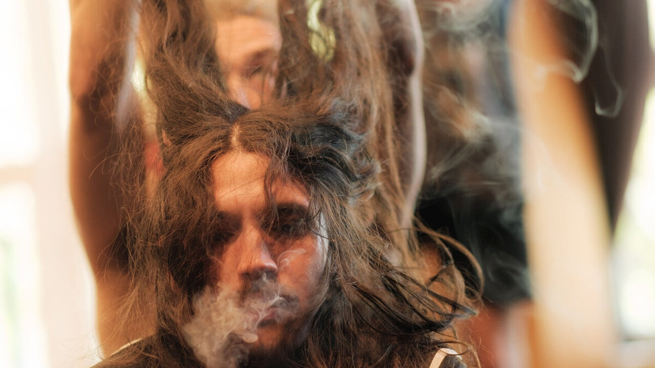 long haired man, exhailing smoke, someone lifts his brown har up in the air. Blurry men in the background.