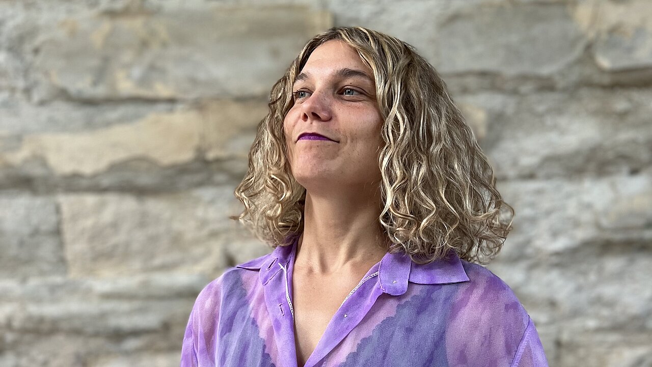 Clara-Marie Müller in a purple shirt in front of a wall made of stone. The chin lifted up, light smile on the face, looking at something in the distance.
