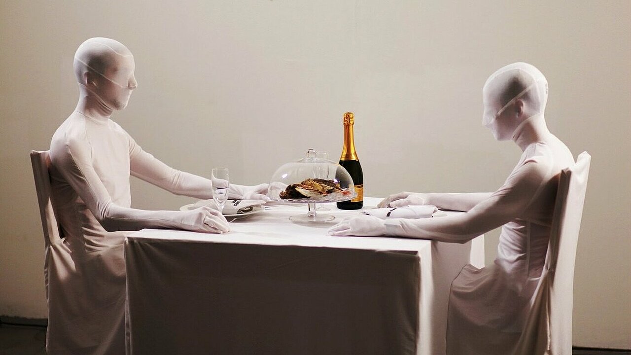 Two people in white fuull body suits sitting at a set dining table.