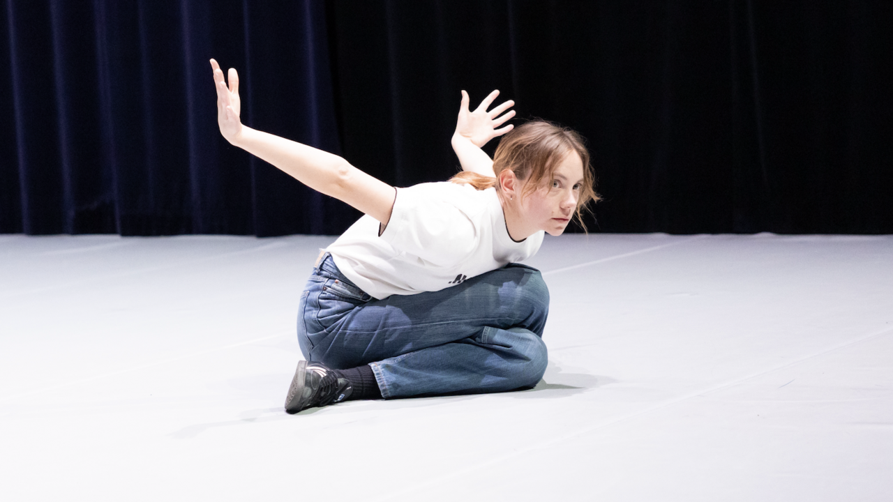 Annabel Koele performing, sitting between her legs, leaning forward, arms in the air
