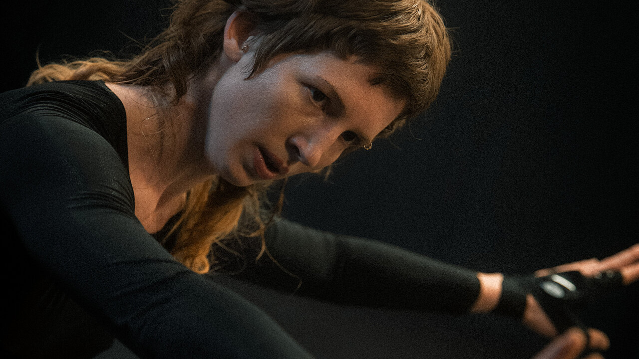 Helén stretching both arms front, performing in a black box, dressed in black
