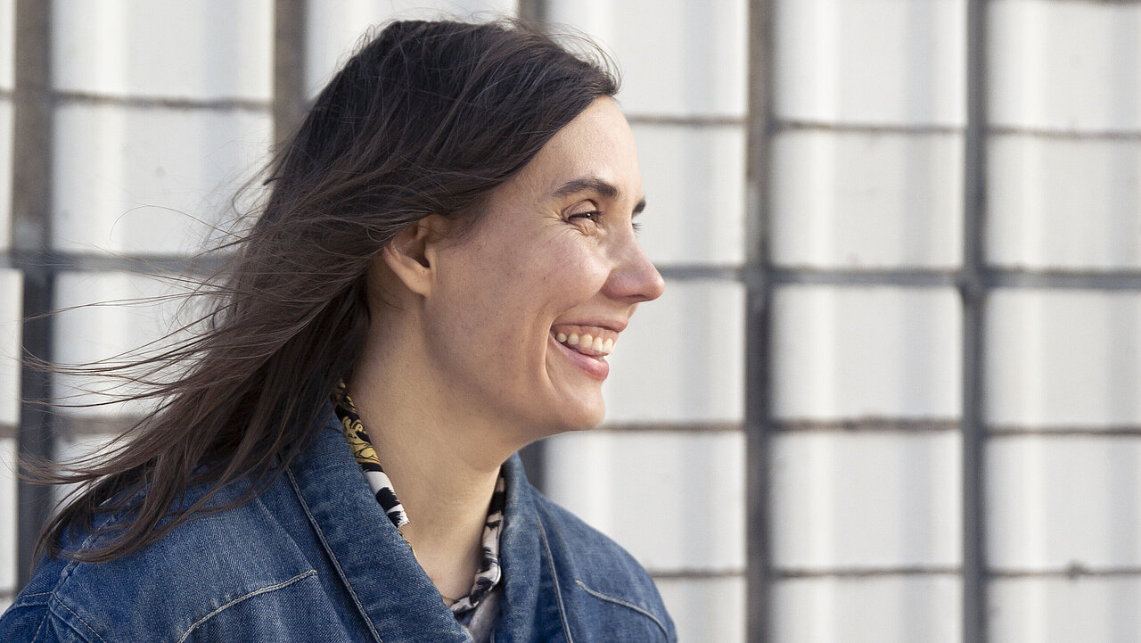 Janina, wearing a jeans shirt, leaning against a bright wall